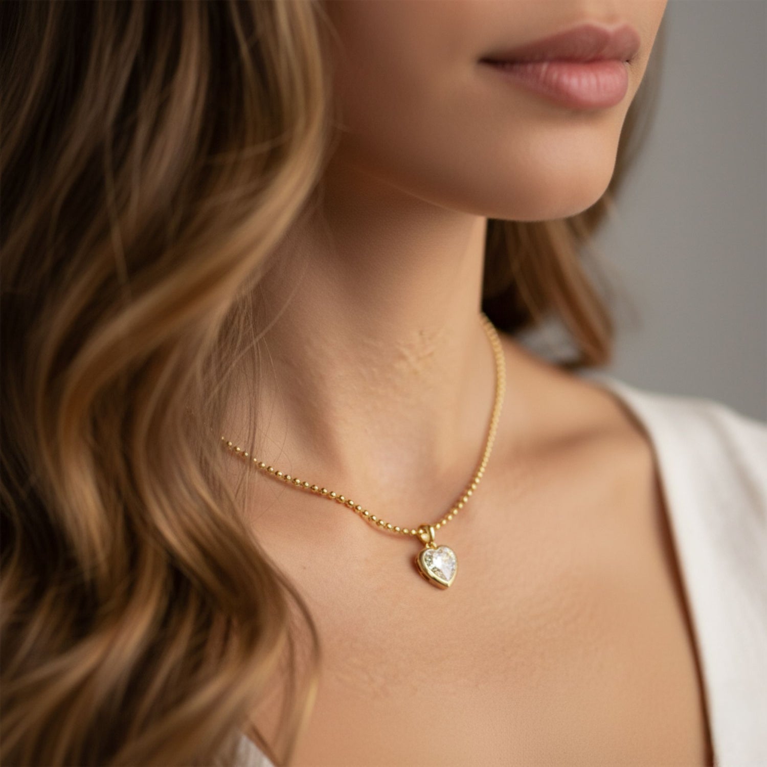 A close-up lifestyle shot of a woman wearing a delicate gold chain necklace with a small heart-shaped pendant featuring a clear, textured inlay.